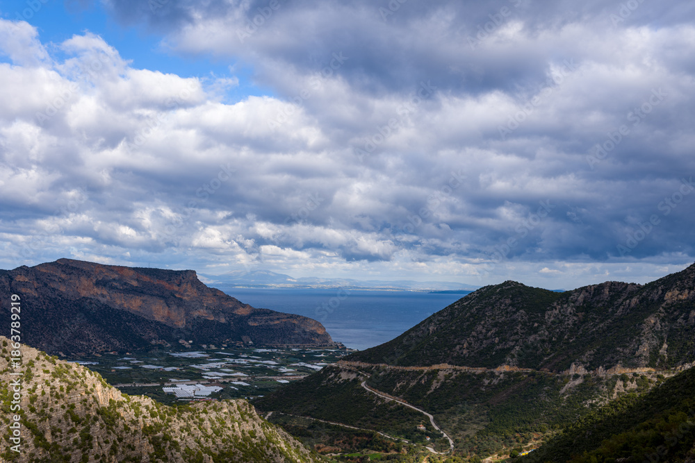 Fototapeta premium Expansive view over a rugged mountain valley descending toward the blue sea, with winding roads and scattered greenery beneath a sky filled with textured, dramatic clouds. The landscape blends rocky