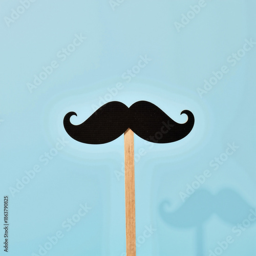 Black mustache on wooden stick against light blue background for Father's Day  