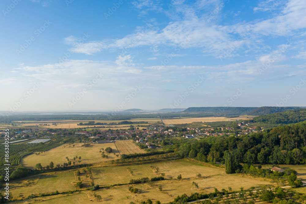 Fototapeta premium Expansive aerial view of Hattonchatel village surrounded by golden farmland, orchards, and dense green woodland under a bright blue sky. The composition highlights the patchwork fields, rural
