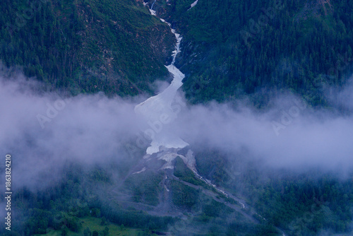 Bild auf Leinwand A winding avalanche chute filled with lingering snow cuts through a lush green mountain valley, partially veiled by low morning clouds and surrounded by dense forest slopes