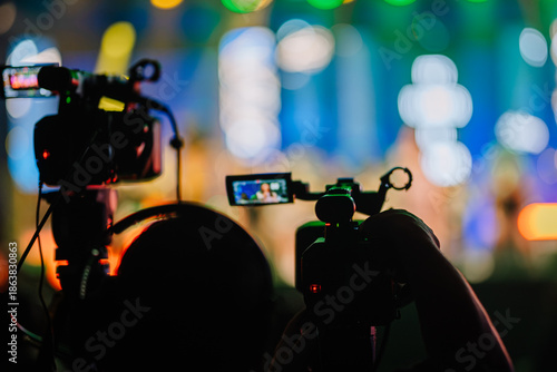 Two video cameras positioned in foreground capturing vibrant live performance on stage, with colorful lights and blurred audience in background, showcasing dynamic event atmosphere