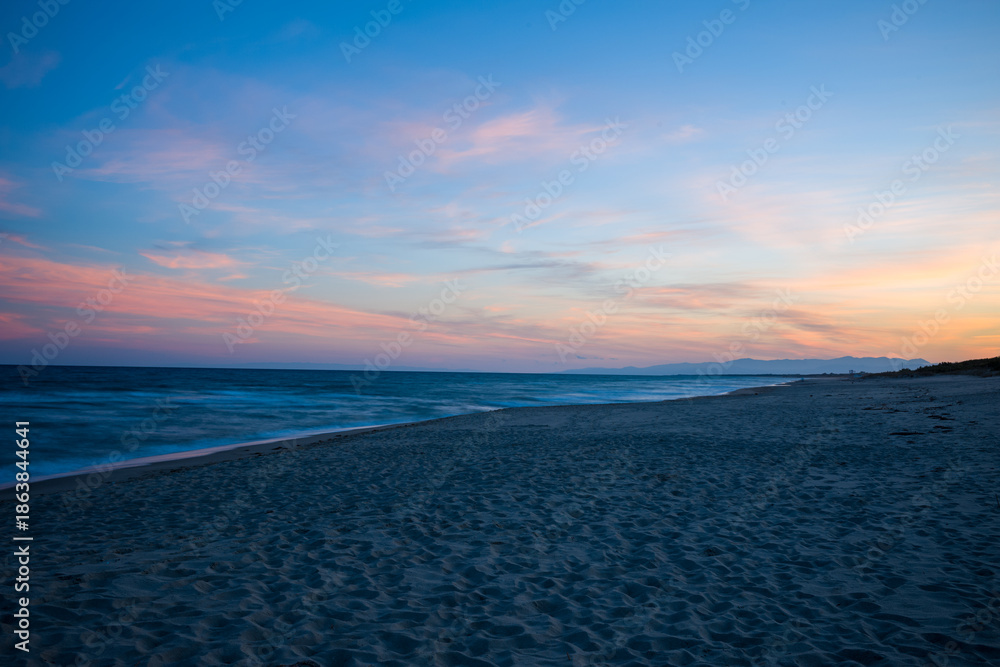 Obraz premium Soft pink and orange hues illuminate the sky above the empty sandy beach at Plage de Acchio, Fiumicello. Gentle waves and distant mountains create a peaceful, expansive seascape in the fading evening