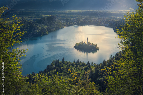 View of an island church rising majestically from a placid lake ringed by verdant forests under a soft, sun-kissed sky, Bled, Radovljica, Slovenia.