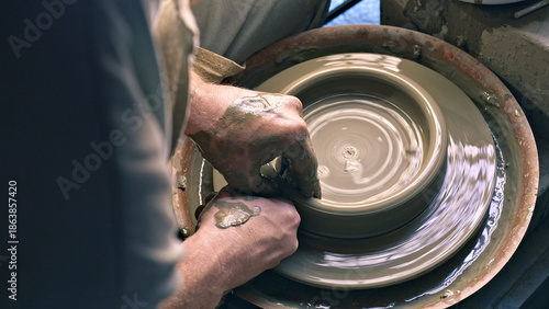 A craftsperson shapes a clay pot on a pottery wheel, their hands skillfully guiding the spinning form. The close-up photo captures a workshop, emphasizing creativity and artistry in pottery making.