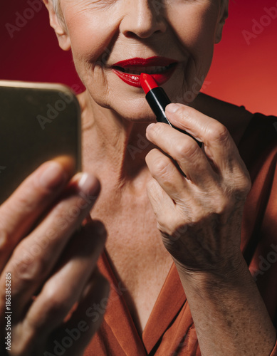 Close-up beauty shot of older woman with wrinkles putting on red lipstick and holding mirror, analog studio shot with film grain structure of female best ager model in her 70s with makeup and red lips