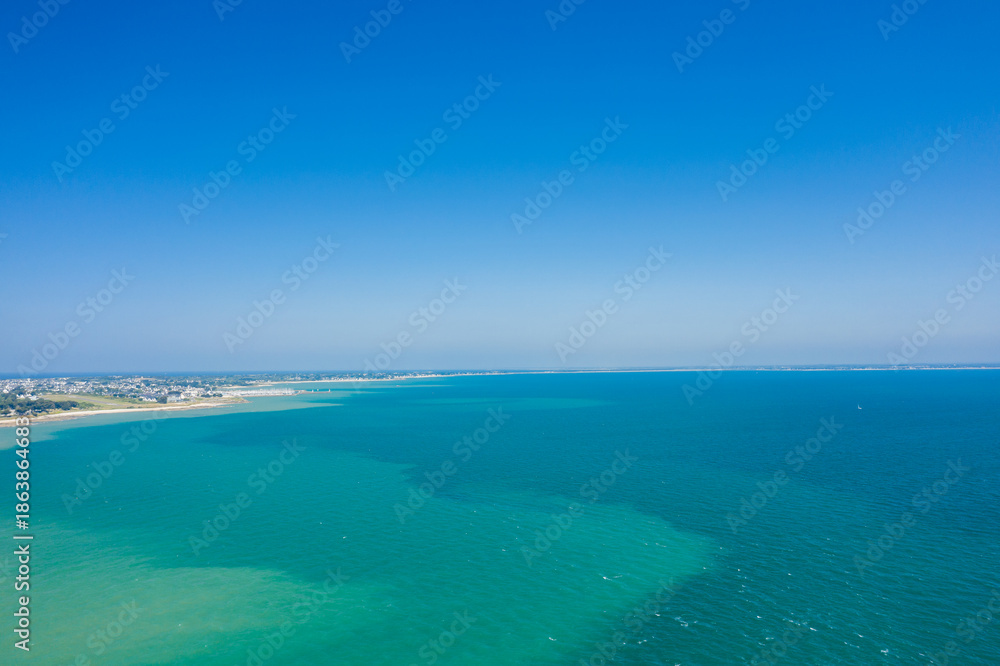 Obraz premium Aerial view of a vast turquoise bay meeting a sandy shoreline near Quiberon, with clear blue skies and distant coastal settlements visible. The scene is sunlit and expansive, evoking a sense of