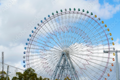 Large colorful Ferris wheel against a blue sky with soft white clouds. Amusement park attraction with multi colored gondolas and steel frame construction. Recreational funfair background.