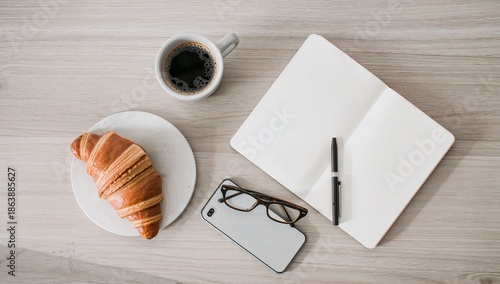 Top view of a light wooden desk with black coffee in a cup a croissant on a plate an open blank notebook with pen eyeglasses and a smartphone arranged neatly for a calm morning workspace
