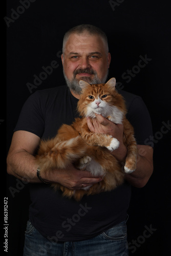 A bearded middle-aged man holding a fluffy ginger cat against a dark studio background..