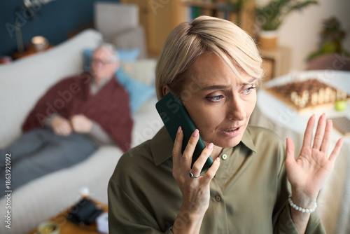 Wallpaper Mural Caucasian middle aged woman talking on smartphone with concerned expression, calling for doctor for sick father, raising hand while sitting in living room, senior man resting on sofa in background Torontodigital.ca