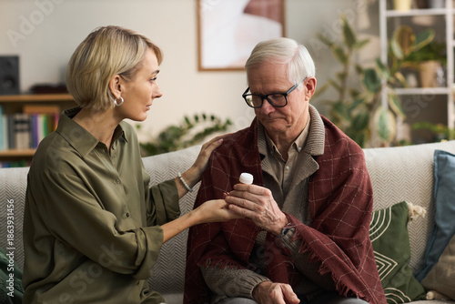 Wallpaper Mural Caucasian middle aged woman supporting senior Caucasian man by handing medication bottle while sitting together on sofa, woman gently touching man's shoulder, plants in background Torontodigital.ca