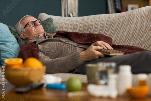 Wallpaper Mural Senior Caucasian man lying on sofa resting with eyes closed, wearing glasses and holding book in hand, medicine bottles and fruit on table in foreground suggesting home recovery Torontodigital.ca