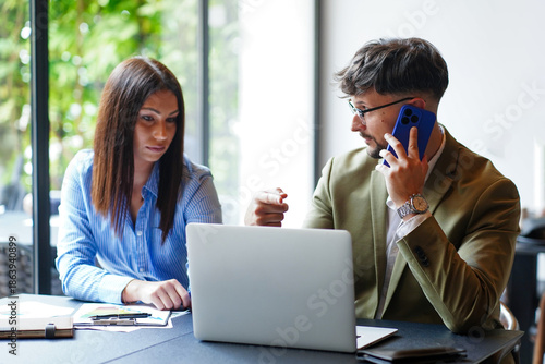 Business meeting in a modern office with a male and female discussing project details while using a laptop and phone