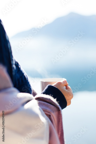 Woman drinking morning coffee in nature outdoors. Traveler relaxing in winter travel, enjoying healthy warm water by mountain lake. Calming lifestyle, slow living. Hand with steaming hot beverage
