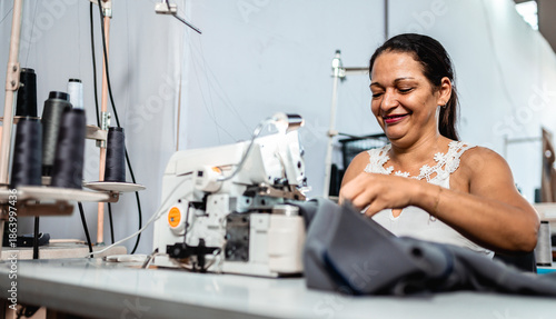 Photos Smiling mature woman working as a seamstress in a garment factory, using an industrial sewing machine to stitch fabric in a bright workshop