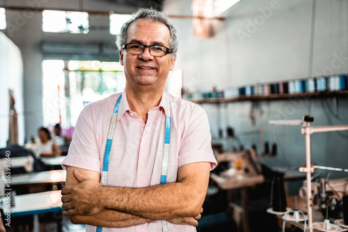 Tableau sur toile Portrait of a confident mature male tailor standing with arms crossed in a garment factory workshop, wearing a measuring tape and smiling at the camera