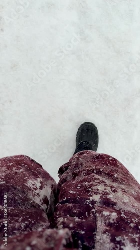 A person is walking in the snow wearing a red coat and black boots. The boots are covered in snow, and the person's feet are visible. The snow is falling lightly, and the person is walking on a pat