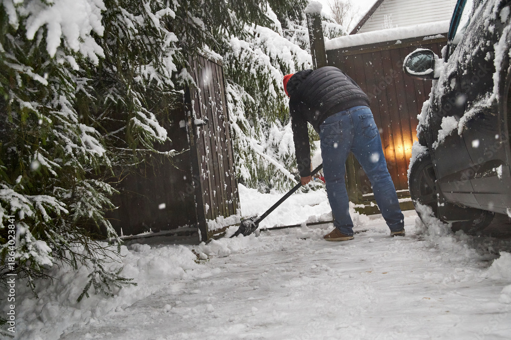 Fototapeta premium Man clearing snow near a car in winter yard, using a snow shovel during snowfall. Outdoor seasonal work, cold weather, driveway cleaning and winter home maintenance