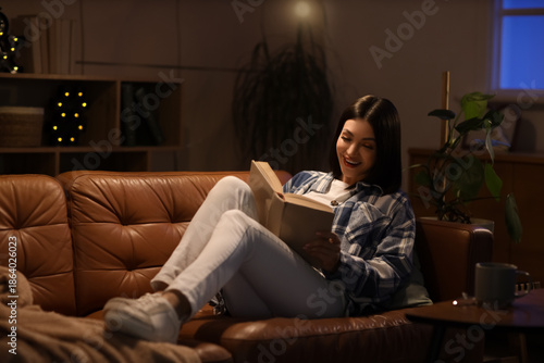 Beautiful young woman reading book and sitting on sofa in living room at night