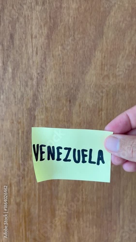 Hand Holding Sticky Note with Venezuela Text over Wooden Background