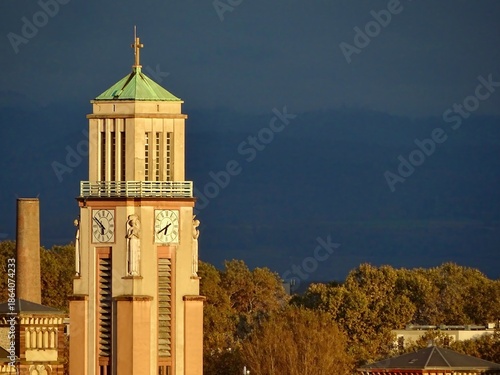 Sainte Jeanne d'Arc Church clock tower in Mulhouse, Alsace, France at golden hour, Art Deco neo-Byzantine brick architecture above autumn rooftops and hills