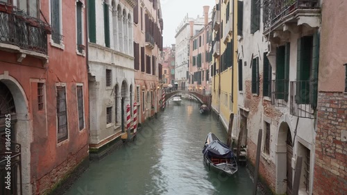 White motorboat navigating narrow Venice canal between historic buildings during rain. Water taxi moving toward camera with moored gondola and people on bridge under umbrellas in Italy.