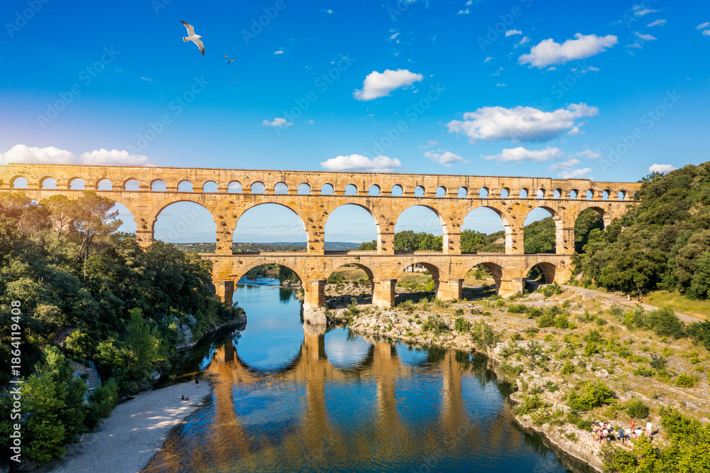 Fototapeta premium Aqueduct of Pont du Gard arches over the river, showcasing the rich history of Vers-Pont-du-Gard, France. Ancient aqueduct of Pont du Gard spans the peaceful river in Vers-Pont-du-Gard, France.