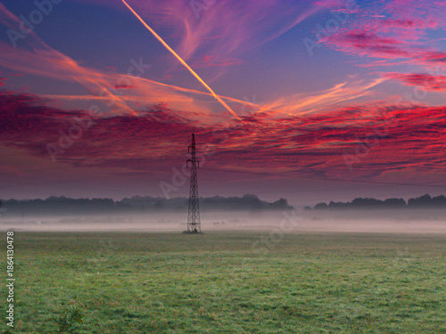 Łąki porośnięte zieloną trawą w letni wieczór. Nad ziemią unosi się warstwa mgły. Niebo jest częściowo pokryte zabarwionymi na czerwono chmurami.