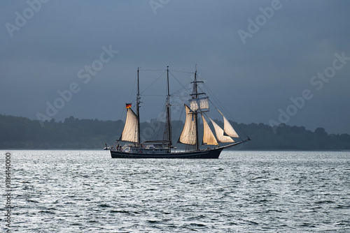A sailing ship with 3 masts and white sails sailing along the coast in bad weather.
