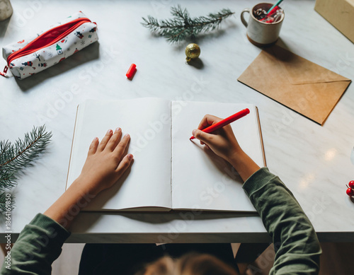 Child writing a letter to Santa in notebook, Christmas desk with decor and gift