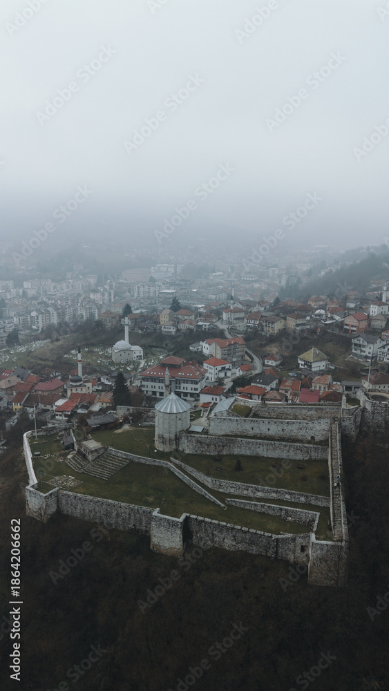 Fototapeta premium Aerial view of Travnik Fortress overlooking the town and surrounding hills, showing historic stone walls, green courtyards and misty mountain landscape from above. Amazing Bosnia and Herzegovina