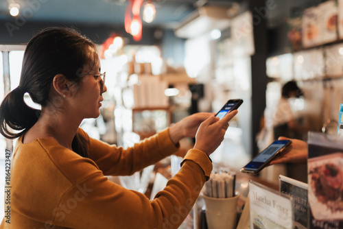 Asian woman customer scanning smartphone to pay at coffee shop counter. Real lifestyle moment for fintech advertising, digital banking apps, QR payment. secure payment messaging, retail technology,
