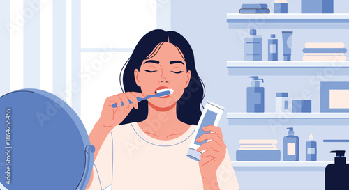 A young woman is standing in her modern bathroom looking in a mirror while brushing her teeth as part of her morning routine.