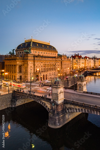 The aerial scene shows the National Theatre in Prague illuminated at dusk. The Smetana embankment and Rasin bridge are visible, creating a lively atmosphere in Czechia.