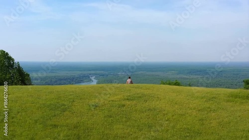 Man sits on hill overlooking river and forest in summer afternoon