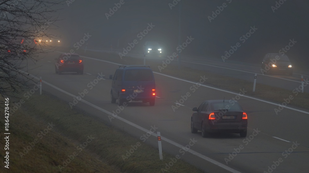 Fototapeta premium Car traveling on the foggy road with headlights or headlamps on. Low visibility - Dangerous driving of cars in winter in bad weather.