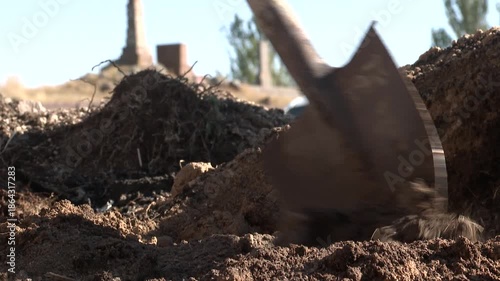 Close up of a gravedigger using a shovel to move soil. The man is filling a grave with earth in a cemetery with tombstones in the background.

