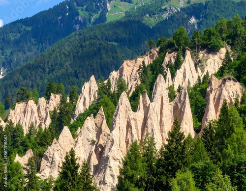 Eroded rock formations, conical in shape, rise amongst evergreen trees against a backdrop of mountains and a blue sky