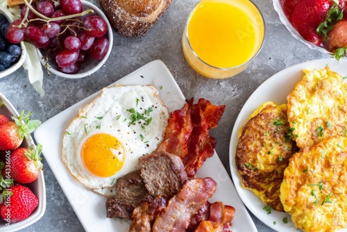 Hearty breakfast spread with fried egg, crisp bacon, savory omelets, fresh fruit, and orange juice flat lay