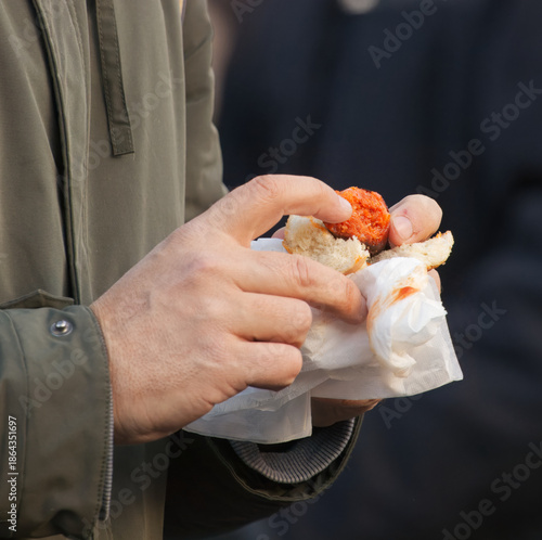 A person in a green jacket holds a small sandwich with red sauce, capturing a casual street meal moment and the simple pleasure of fast food enjoyed outdoors at Naplavka street farmers market.