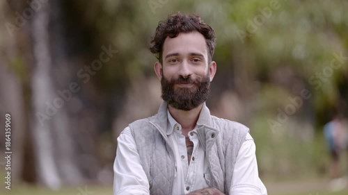 Portrait of a happy young man with a beard and curly hair smiling at the camera in an outdoor park setting.