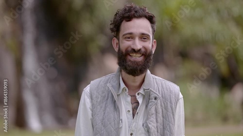 Portrait of a happy young man with a beard and curly hair smiling at the camera in an outdoor park setting.