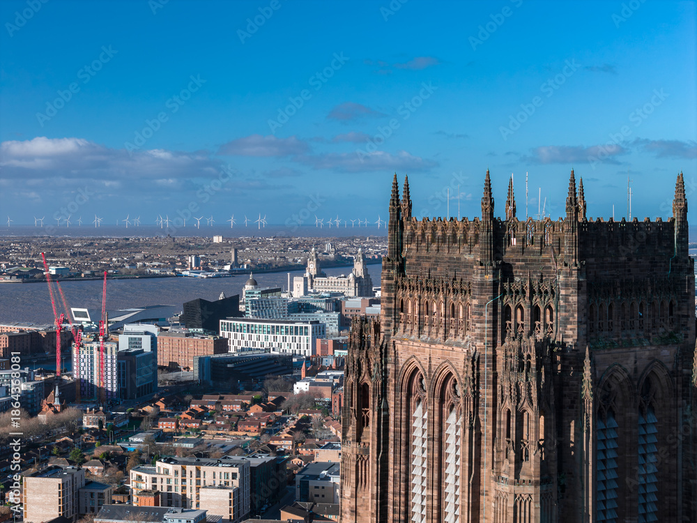 Fototapeta premium Aerial sunrise view of Liverpool shows Liverpool Cathedral, Royal Liver Building, Albert Dock museums, red cranes, modern blocks, and offshore wind turbines.