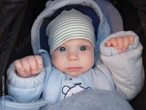 Quadro em tela A baby with blue eyes wearing a light blue outfit and a striped hat is sitting in a stroller