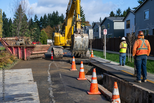 Excavator bucket attachment removing section of asphalt cut in road to dig a utilities trench, public infrastructure construction site, workman watching the project
