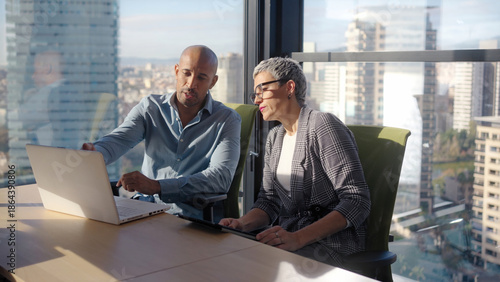 A male and female coworker analyze work on a laptop beside a large window with skyline views, highlighting problem solving, coordination, and an efficient workflow in a contemporary office.