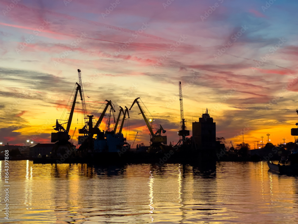Obraz premium Sunset over a busy port with cranes reflecting in the water during evening hours