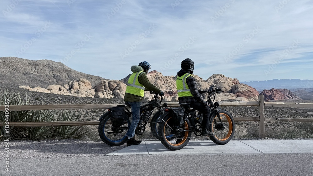 Obraz premium 🚲Back view of cyclists in yellow vests and helmets on e-bikes by a wooden fence. They are looking at the vast Red Rock Canyon desert landscape. Sunny day, travel and active lifestyle concept.