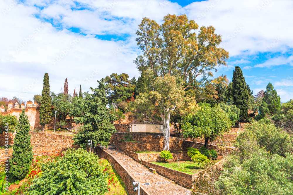 Naklejka premium Castillo de Gibralfaro in Malaga, Spain, showcasing stone walls and lush gardens under sky. Historical fortifications and abundant Mediterranean vegetation on the hill overlooking the city