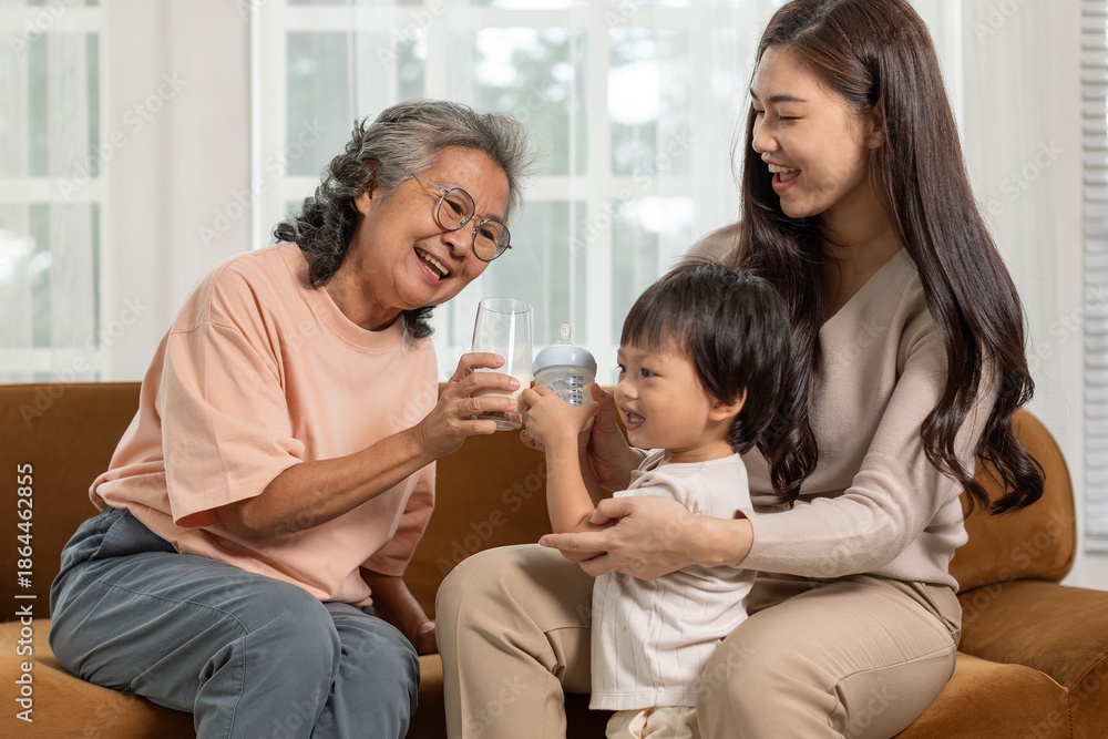 Fototapeta premium Three generations of happy asian family relaxing on brown sofa at home featuring smiling grandmother giving water to cute grandson held by cheerful mother bonding together with love care and joy